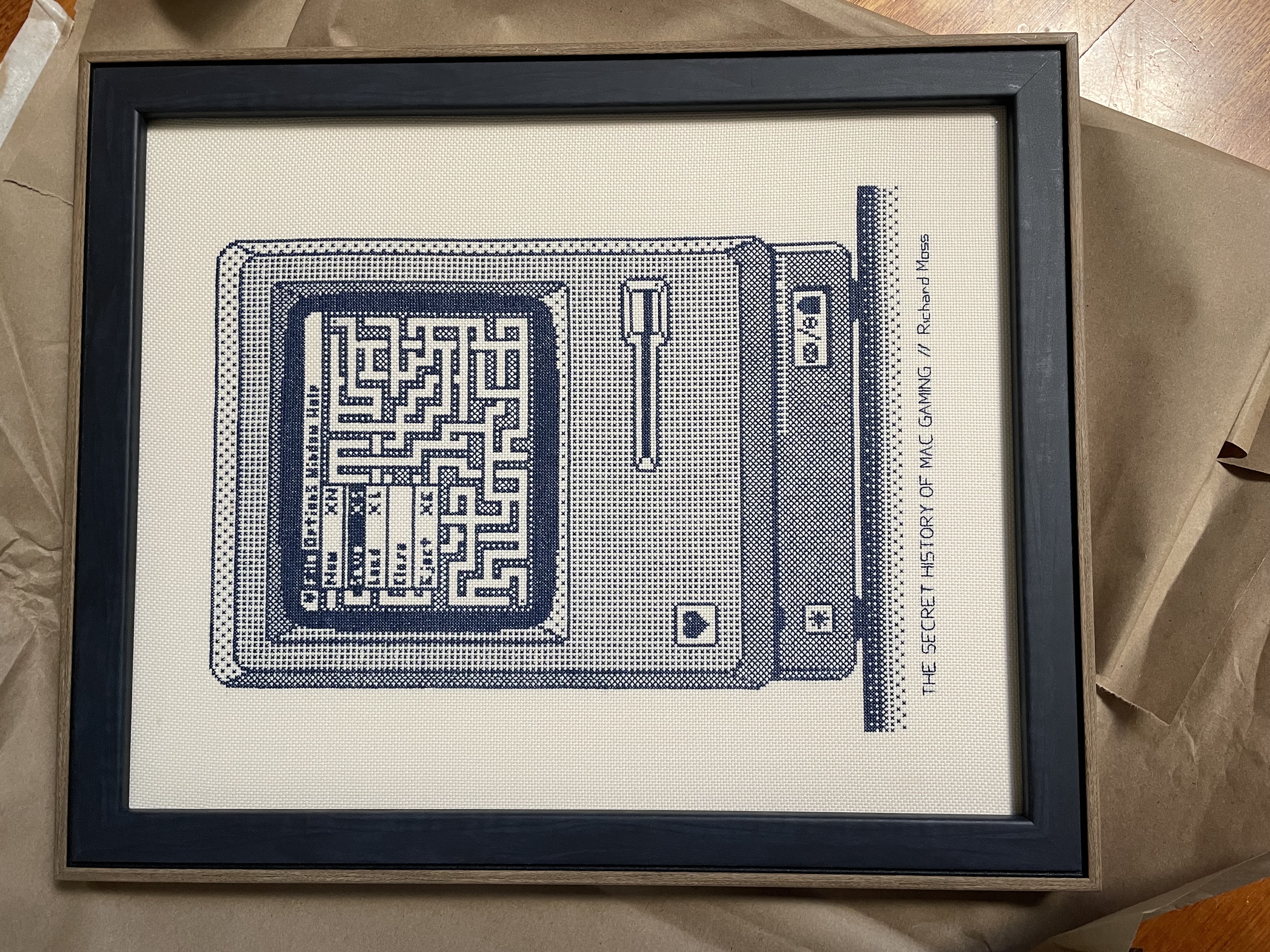 Photo of a framed cross stitch depicting the Secret History of Mac Gaming book cover. It's an illustration of a Macintosh Plus with the game Amazing! on the screen, with a pull down menu open, and the book title and author name underneath.