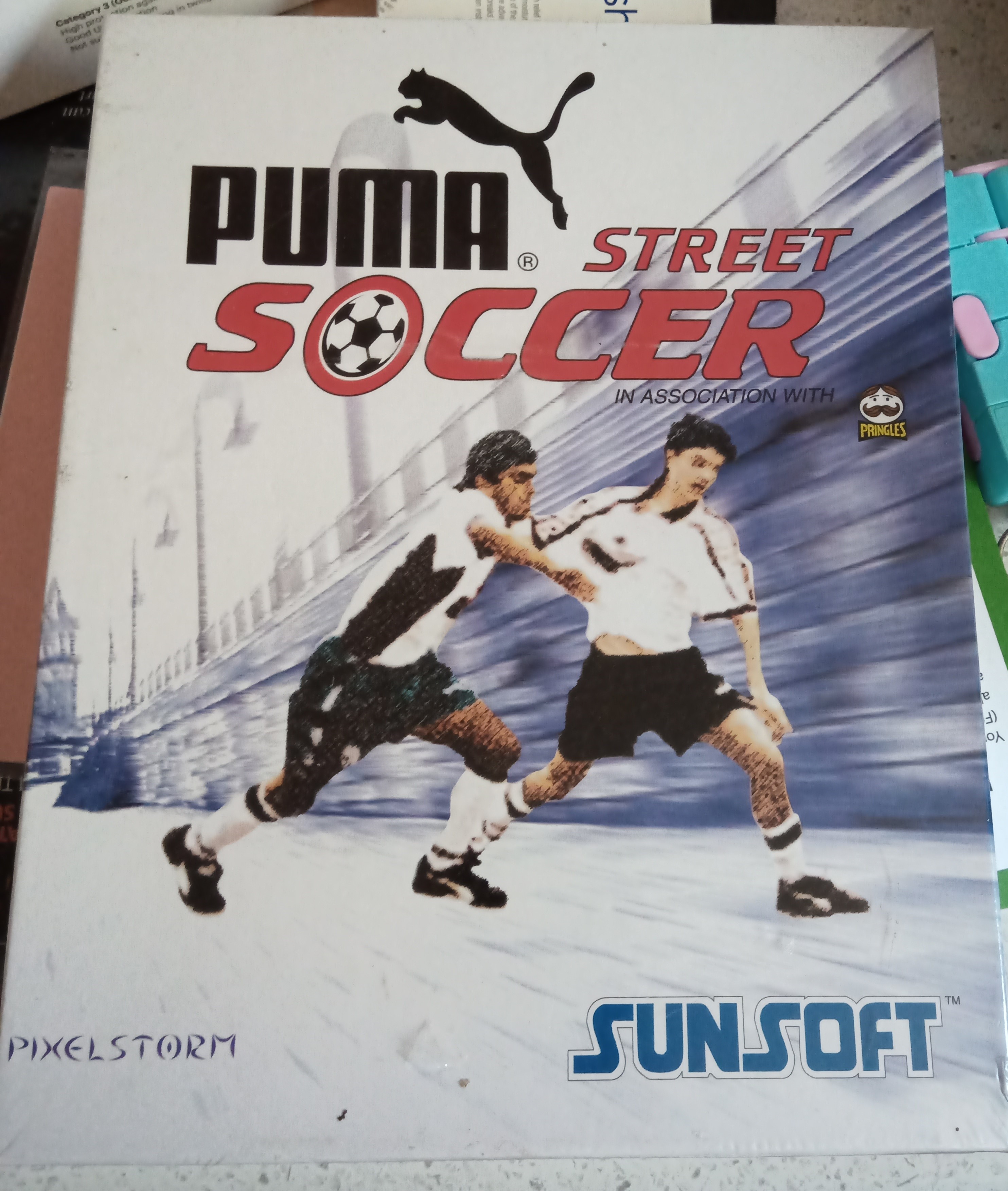 Front cover for the PC version of Puma Street Soccer, showing two players in full kit battling for control of the ball while they apparently rush past some sort of old wall lined by lampposts.