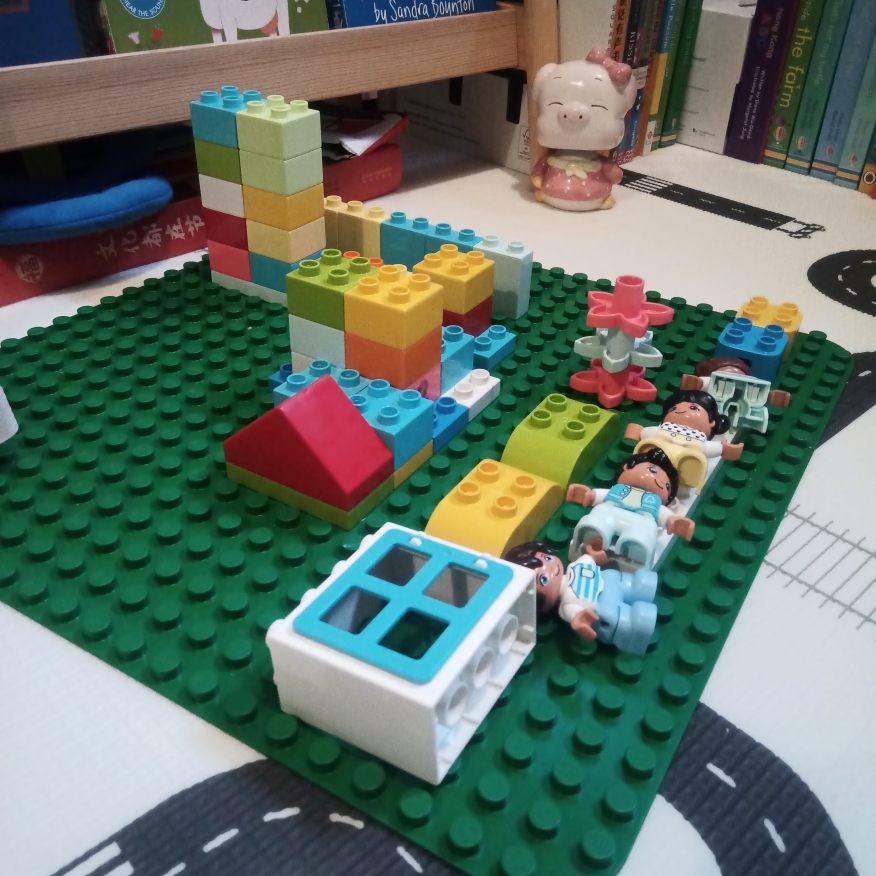 A Lego Duplo town made from a variety of different piece types, with four Duplo people laying down on a bench/bed near one edge.